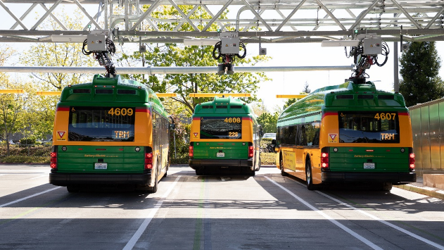 Three green and yellow buses are parked under a charging station, with trees in the background.